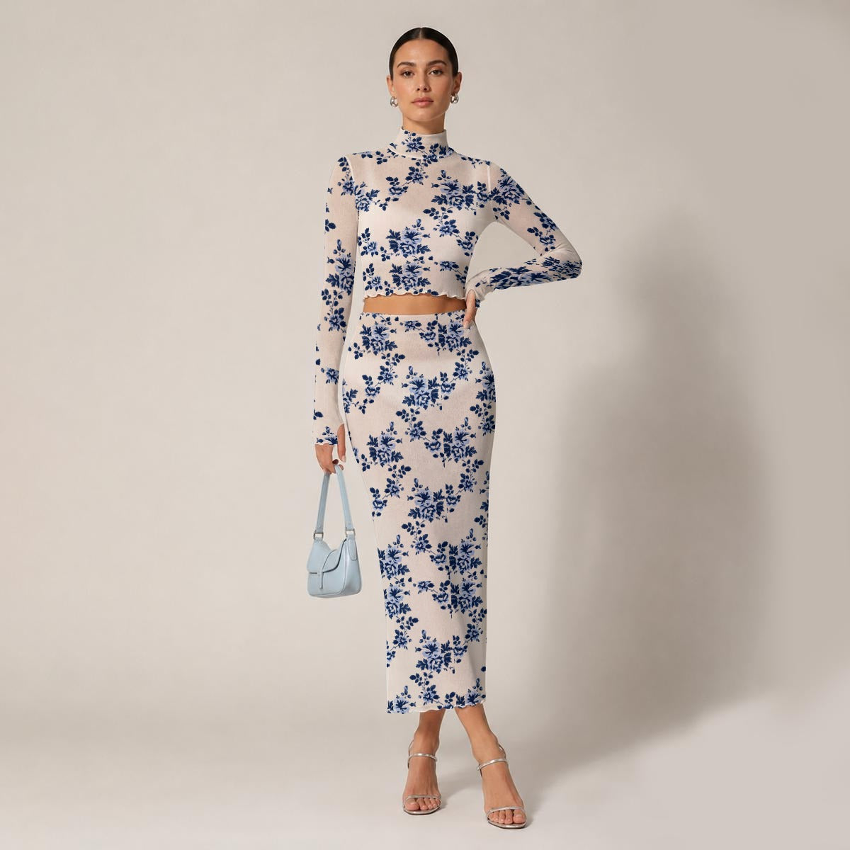A model stands against a light beige studio background wearing a sheer long-sleeve mock-neck top and matching midi skirt set in an off-white fabric with a blue floral pattern. She is holding a small light blue handbag and wearing clear high-heeled sandals.