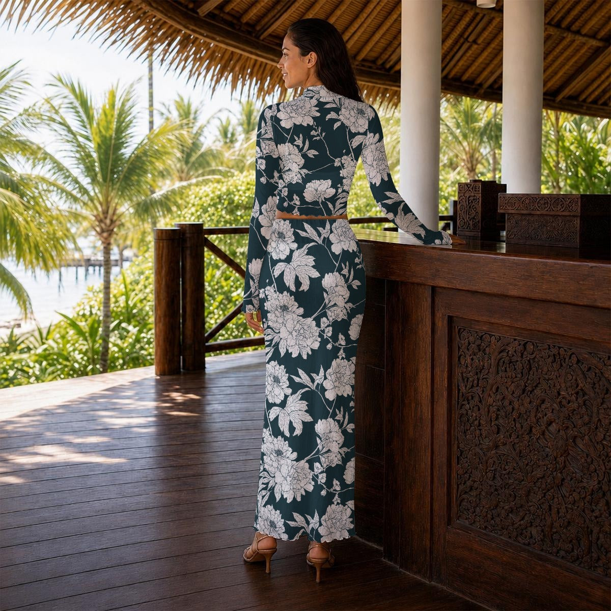 A woman stands on a tropical wooden deck wearing a long-sleeve maxi dress with a dark gray and white floral pattern. She is photographed from behind, looking out over a palm tree-lined ocean view under a thatched roof.