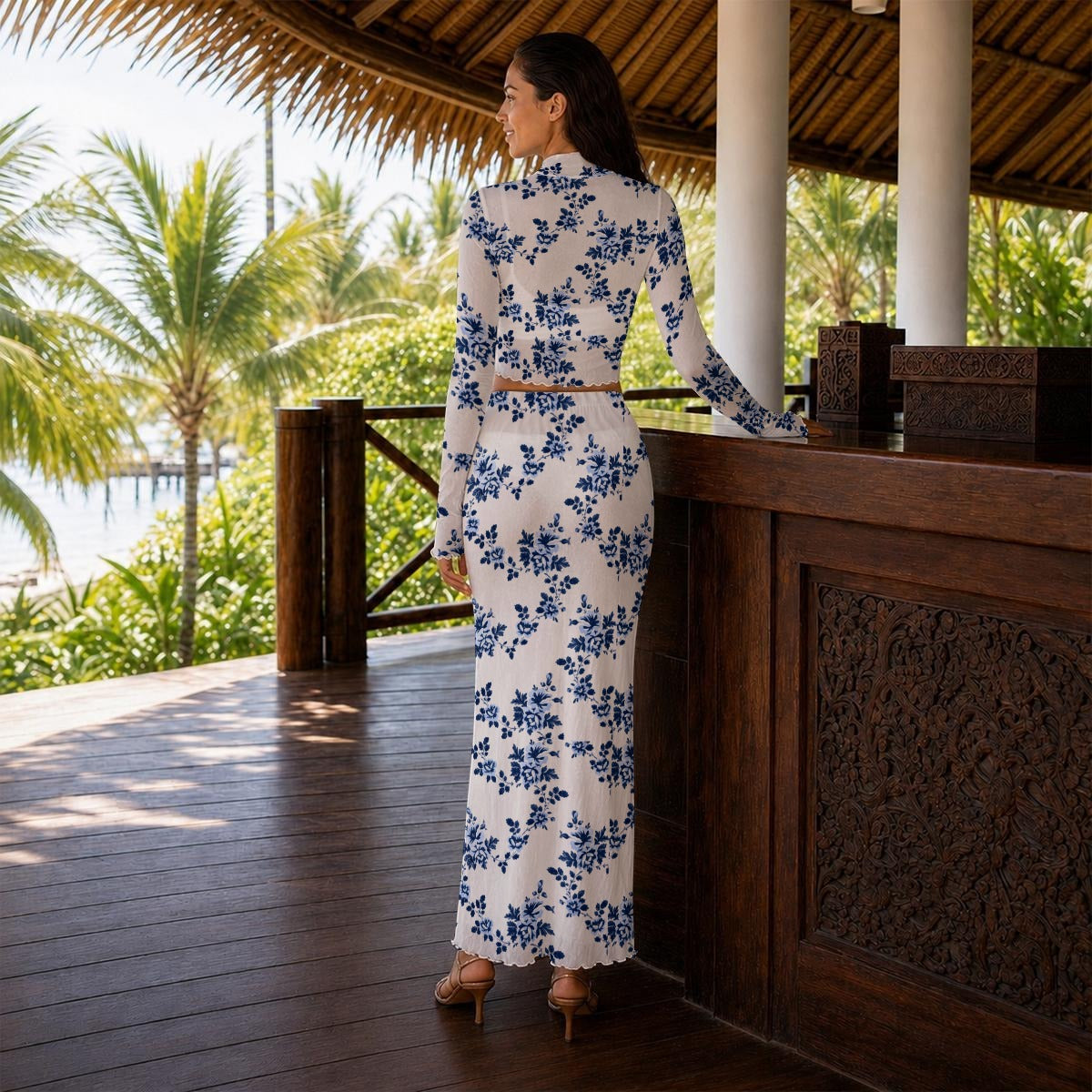 A woman stands on a wooden deck under a thatched roof, wearing a white long-sleeve maxi dress with a blue floral pattern. The setting features palm trees, water, and a tropical resort-style environment.